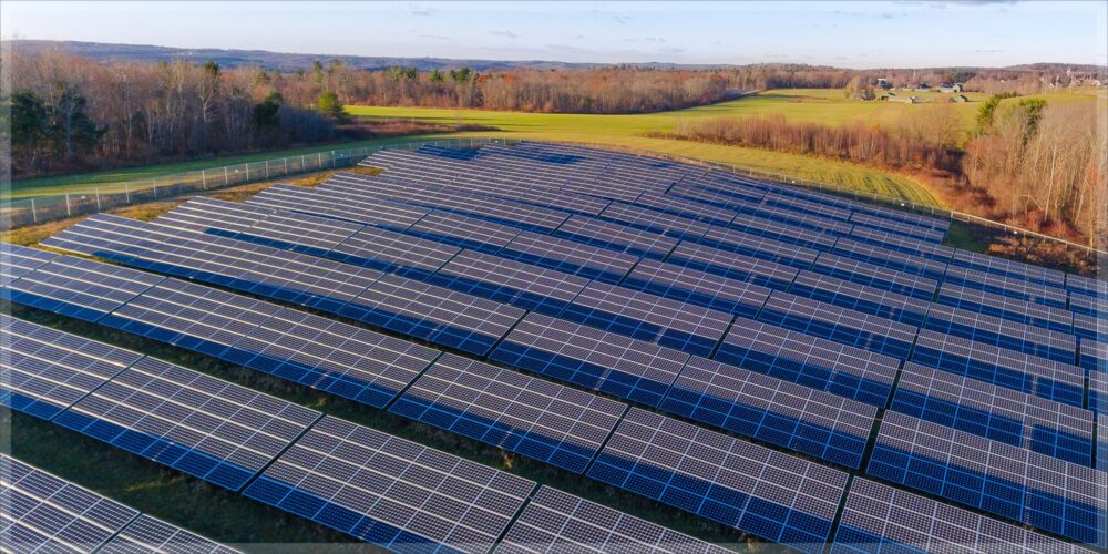 Aerial view of a large solar panel array installed in a field, surrounded by trees and open landscape under a clear sky.