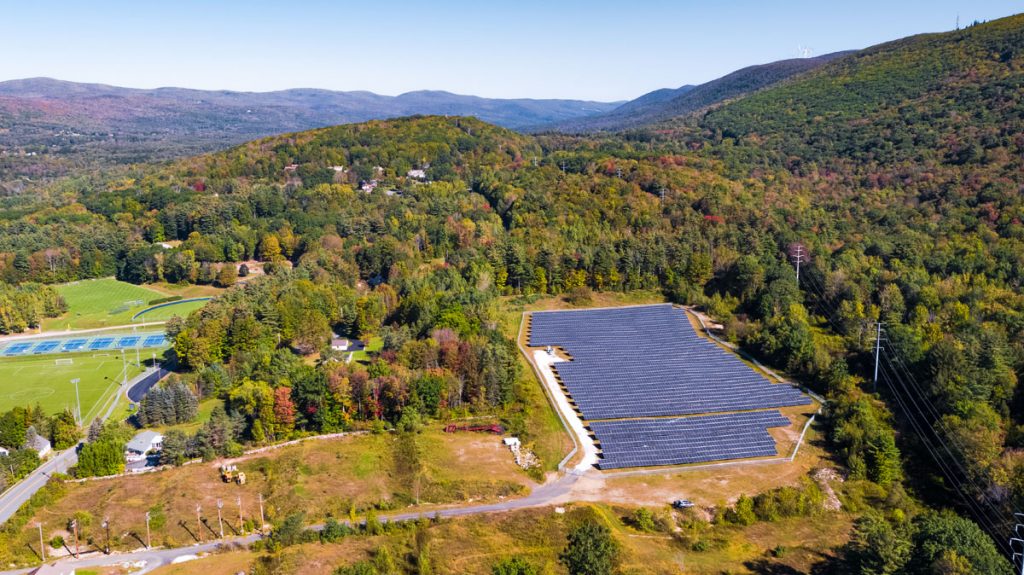 A bird's eye view of a community solar garden surrounded by trees and power lines