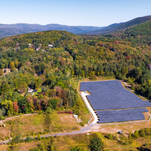 A bird's eye view of a community solar garden surrounded by trees and power lines