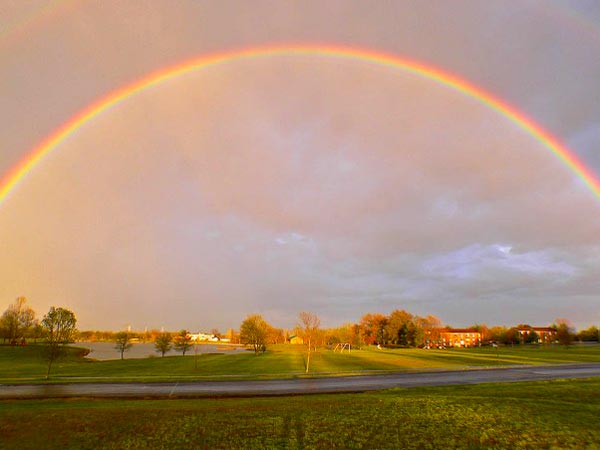 A bright rainbow in a cloudy sky over a suburban neighborhood.