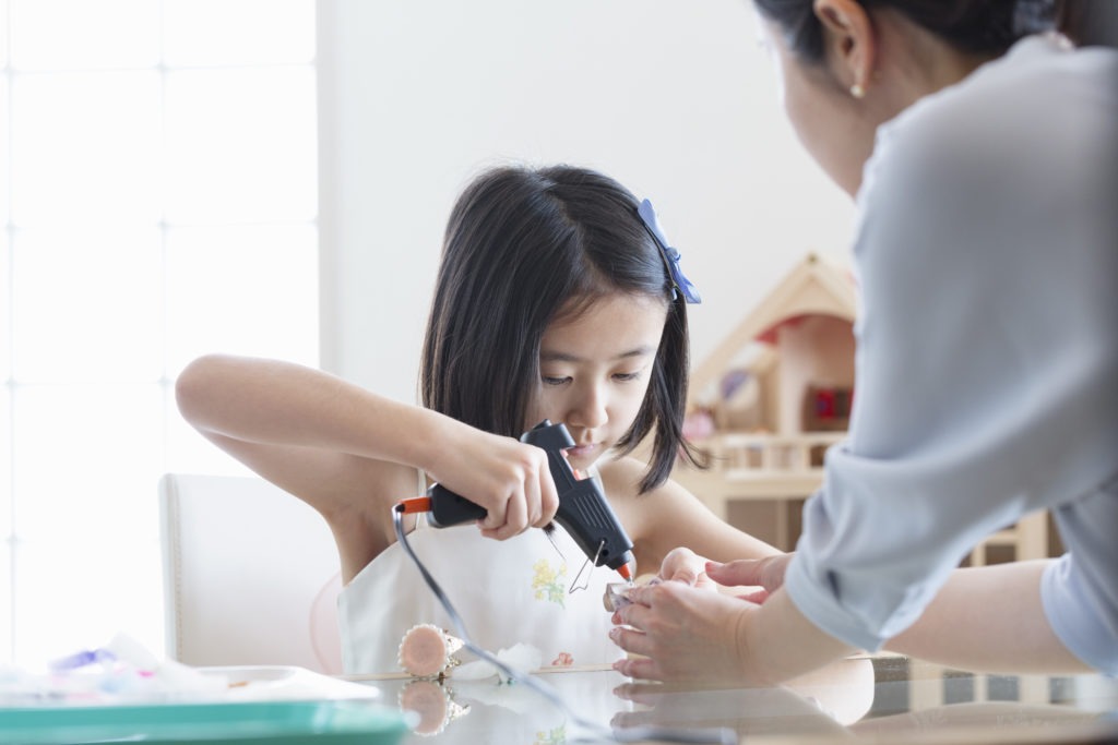 A parent assisting a child with using a hot glue gun