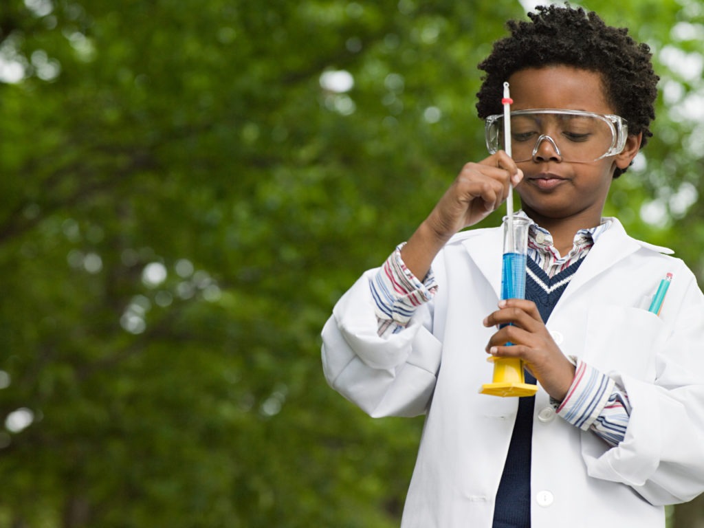 A child in a lab coat sticking a thermometer into a graduated cylinder filled with blue liquid