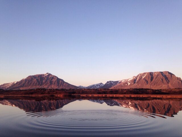 Two mountains are reflected in a calm lake at sunset, with gentle ripples spreading across the water in the foreground.