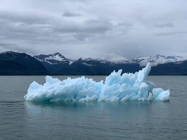 A jagged blue iceberg floats in calm water with snow-capped mountains and a cloudy sky in the background.