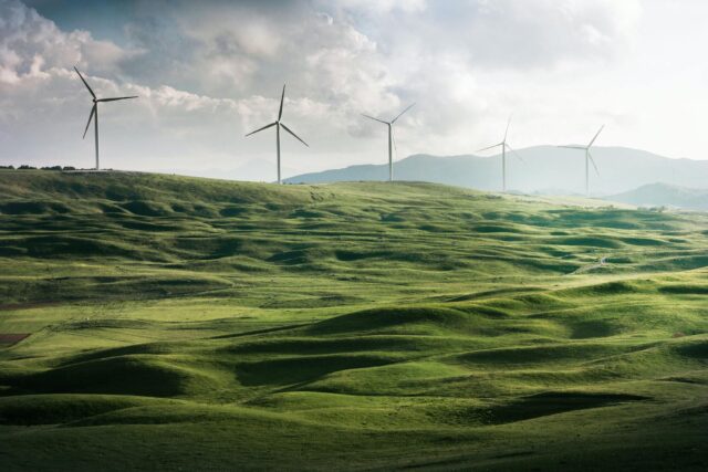 Wind turbines stand on rolling green hills under a cloudy sky, with mountains visible in the background.