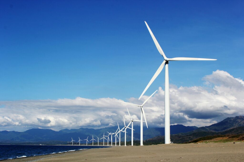 A line of white wind turbines on a sandy beach near a body of water.