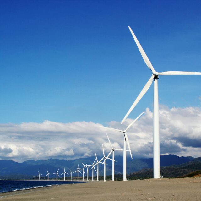 A line of white wind turbines on a sandy beach near a body of water.